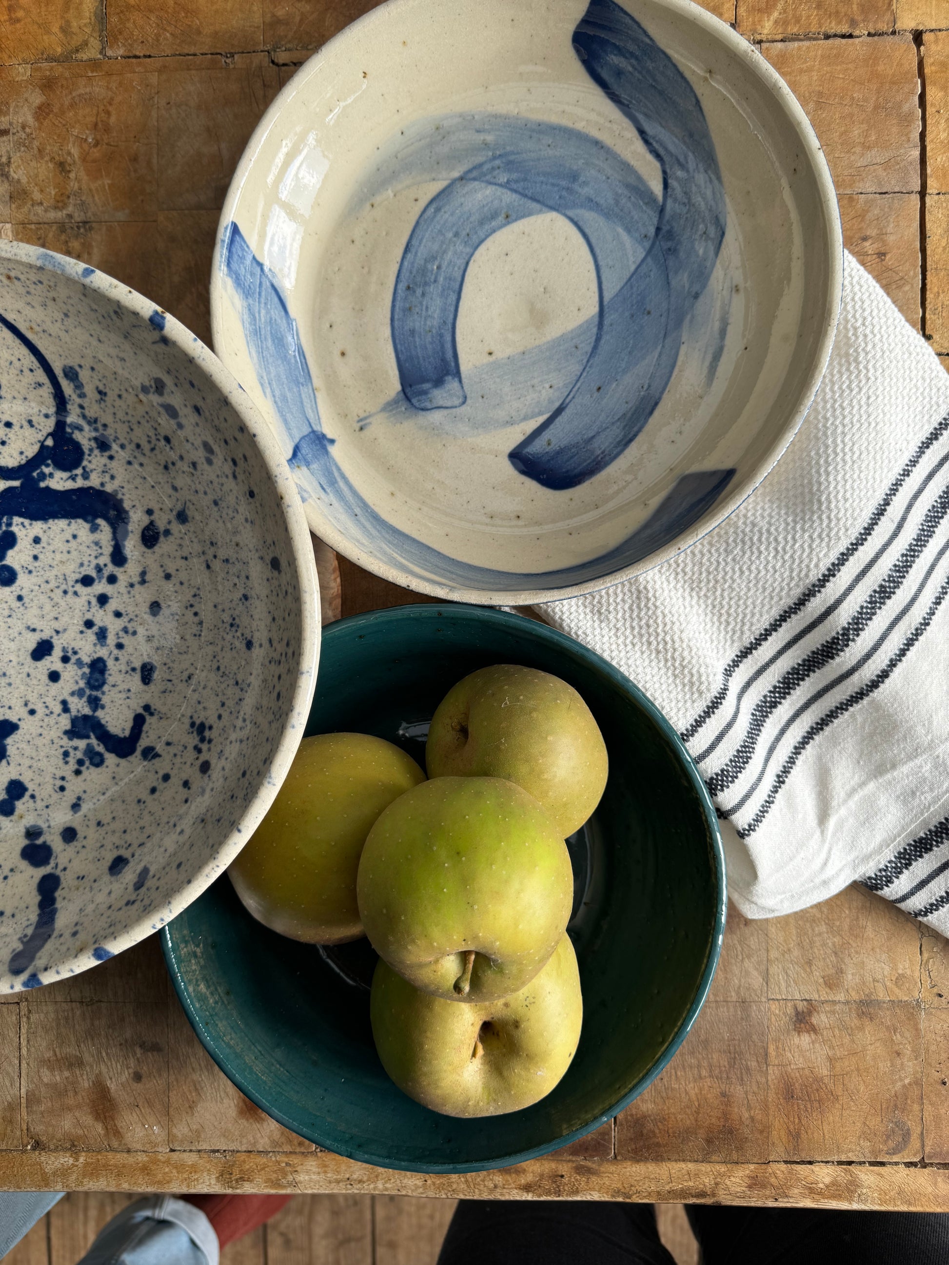 Photo looking directly down on three handmade bowls by Herridge Pottery on a table. One of the bowls contains 4 apples. There is also a tea towel to the side. The bowls are all different colours and sizes, the largest is blue splatter, middle is blue swish and the smallest, with the apples is Malard green.