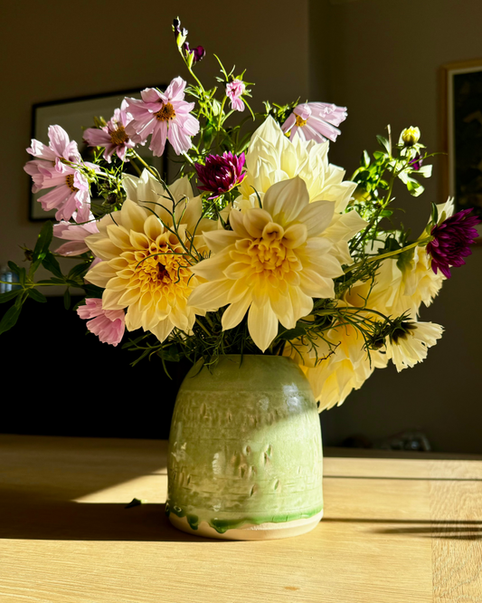 Handmade ceramic green vase where the glaze has dripped down. The vase contain a bunch of dahlias and cosmos.