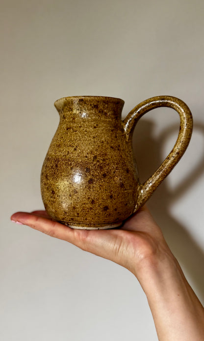 Hand holding a brown ceramic pitcher against a plain background