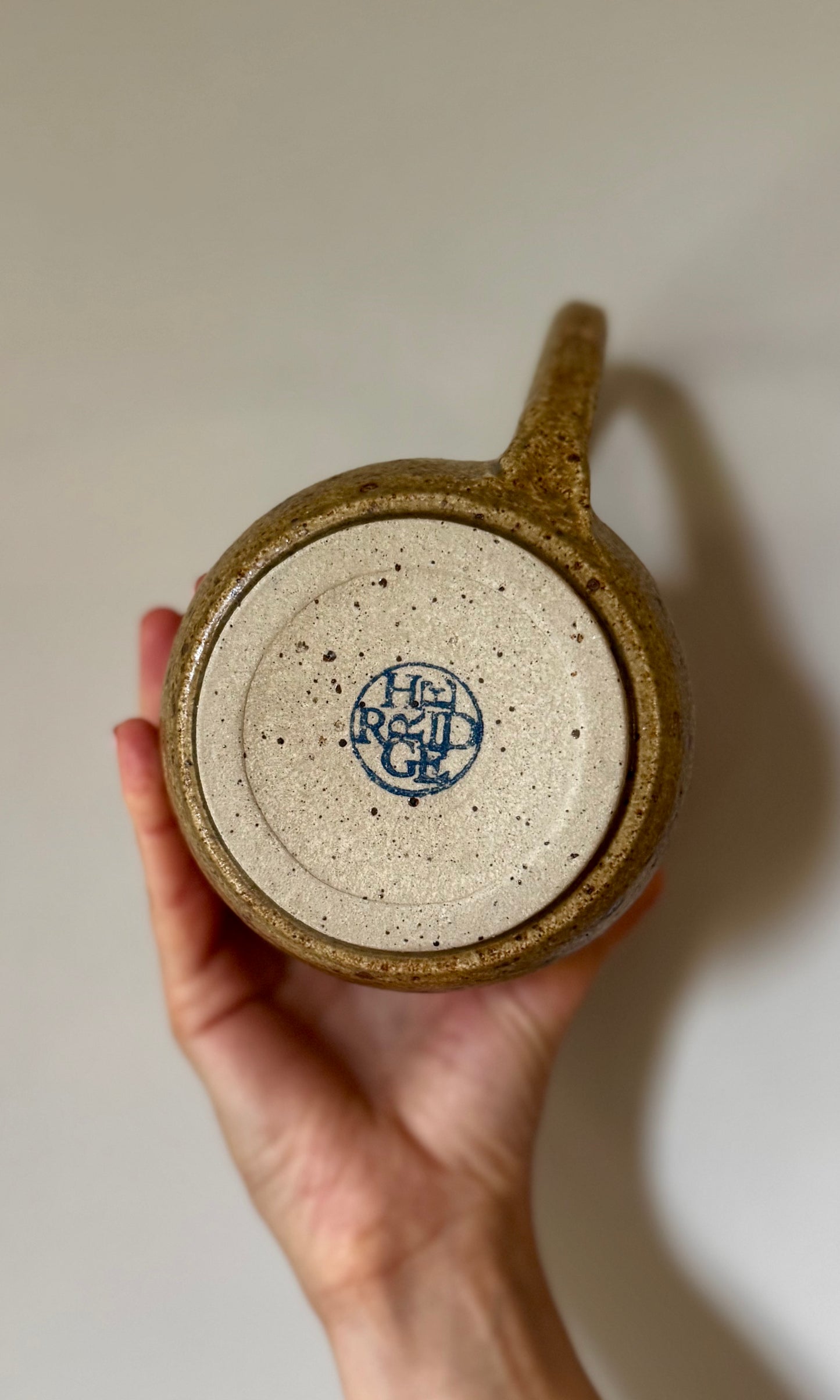 Hand holding a ceramic jug with a visible Herridge logo on a plain background