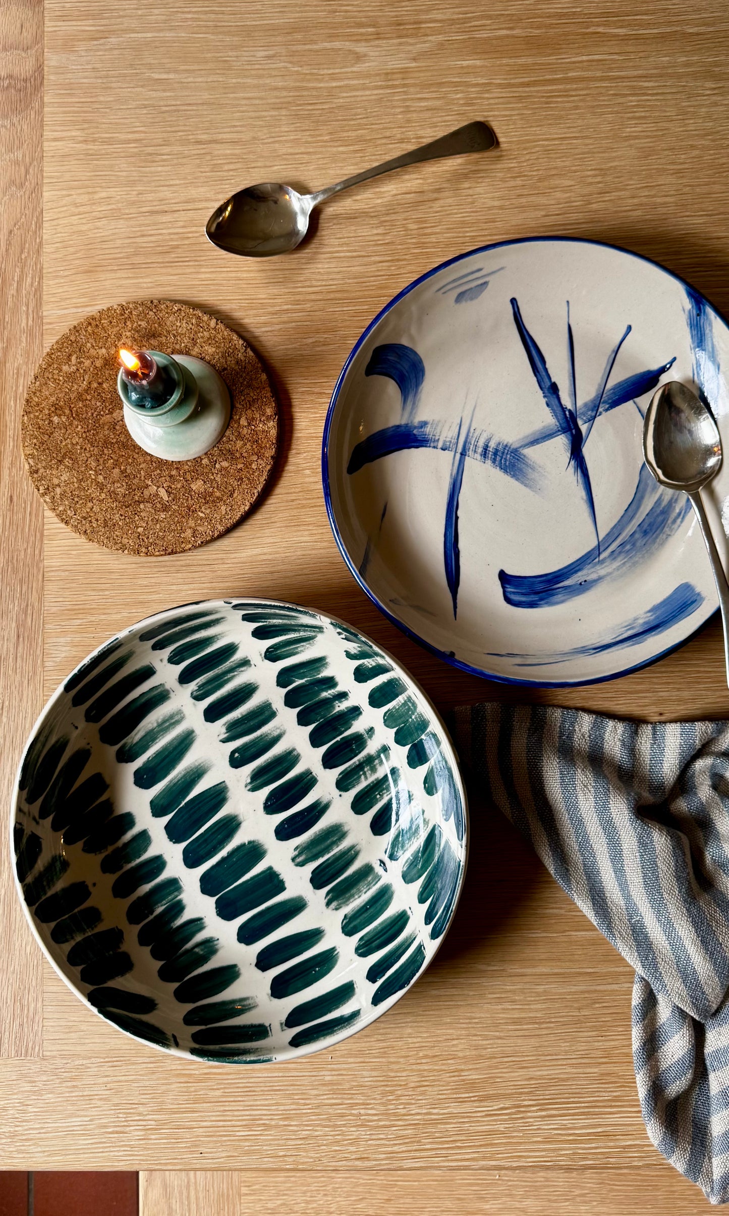 Two ceramic bowls with abstract designs on a wooden table with a candle and spoons.