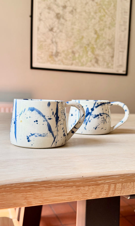 Two white ceramic mugs with blue paint splatter patterns resting on a light wood table, with a framed map hanging on the wall in the background.